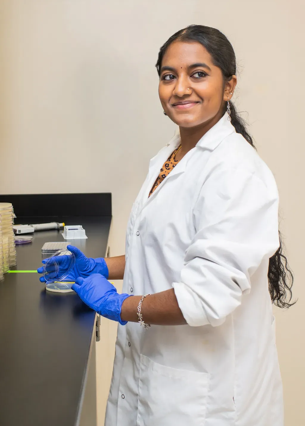 A UNE student in a white coat holds the lid to a petri dish at the counter of ther Burkholder lab while smiling toward the camera