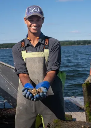 Michael Scannell in waders and a U N E baseball cap holds oysters in his hands while out on a boat