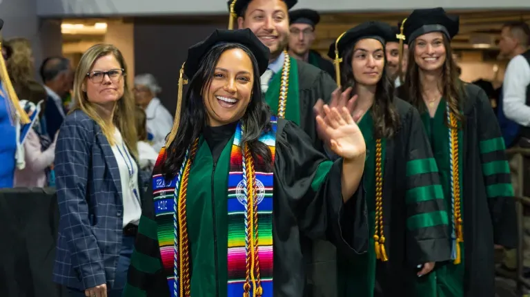 A UNE graduate wearing doctoral regalia waves walking into the ceremony