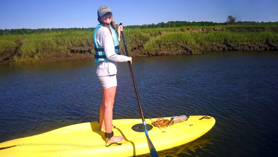 A student smiles at the camera while paddle boarding