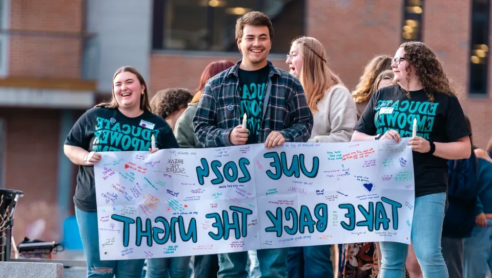 Three students hold up a banner that reads "UNE 2024 Take Back the Night"